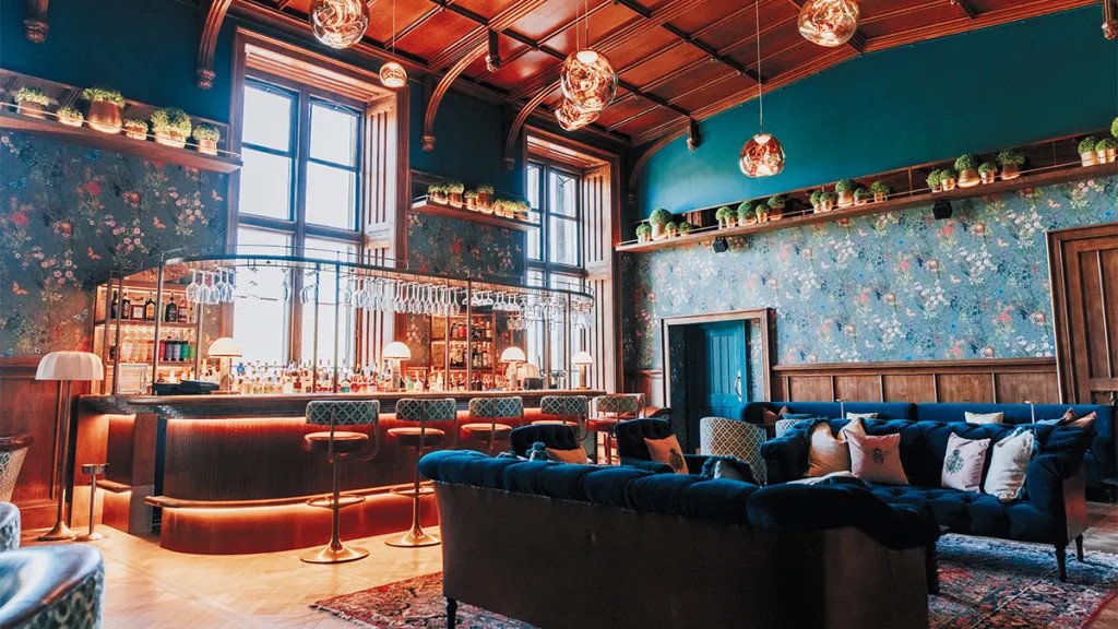 The bar at Mar Hall: a panelled room with a lit bar, blue velvet sofas, floral walls and copper pendant lights